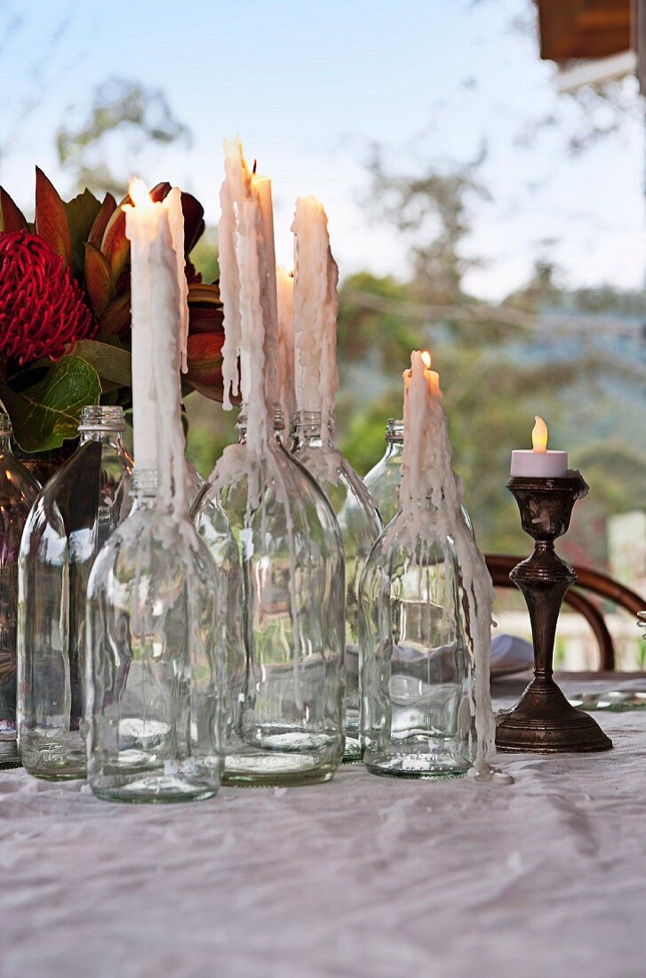 Lit, dribbling candles in empty bottles and silver candlestick on table in garden