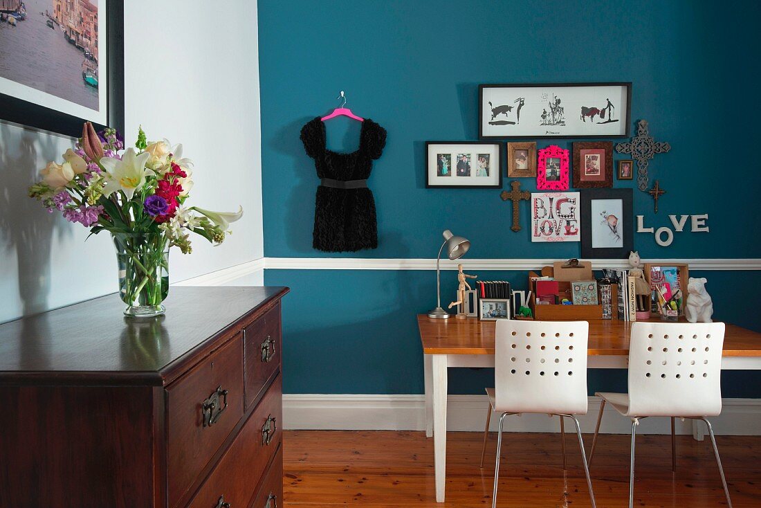 Schreibtisch mit zwei Stühlen vor blauer Wand; Blumenstrauss auf traditioneller Kommode im Vordergrund