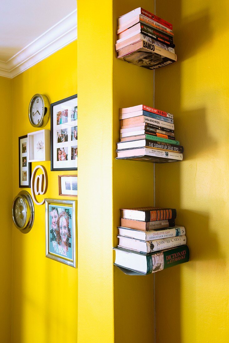 Pictures and clocks on yellow wall separated from floating bookshelves in niche by pillar