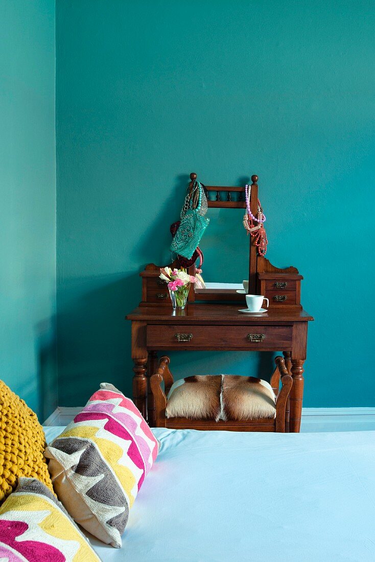 Bed with colourful scatter cushions and antique dressing table and stool in turquoise bedroom