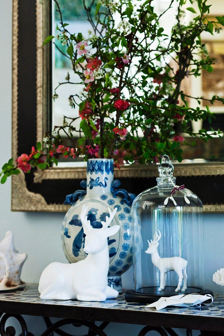 Hirschfiguren aus weißem Porzellan und Keramikvase mit blühenden Zweigen vor elegantem Spiegel