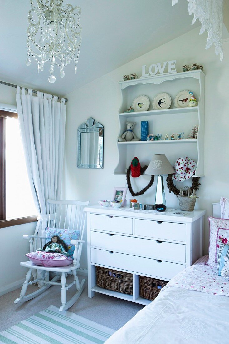 Kommode und Wandregal mit romantischer Deko und Puppe auf Schaukelstuhl in weißem Kinderzimmer