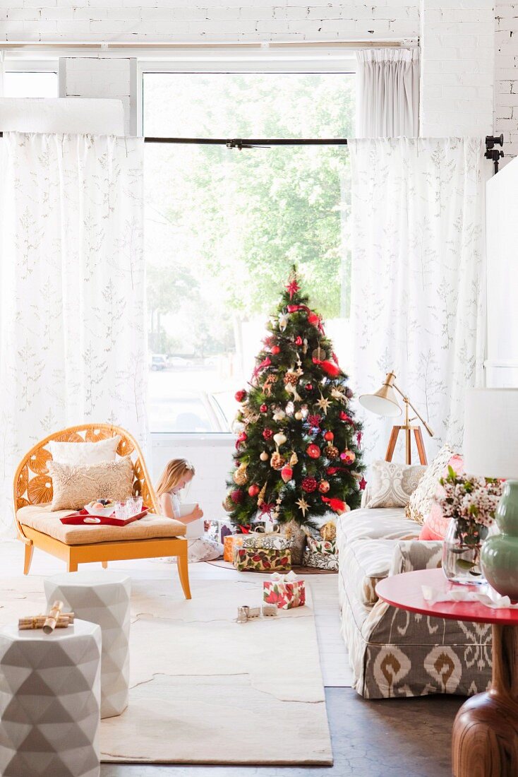 Wohnzimmer mit Sessel und Sofa, im Hintergrund Mädchen vor Weihnachtsbaum mit Geschenken