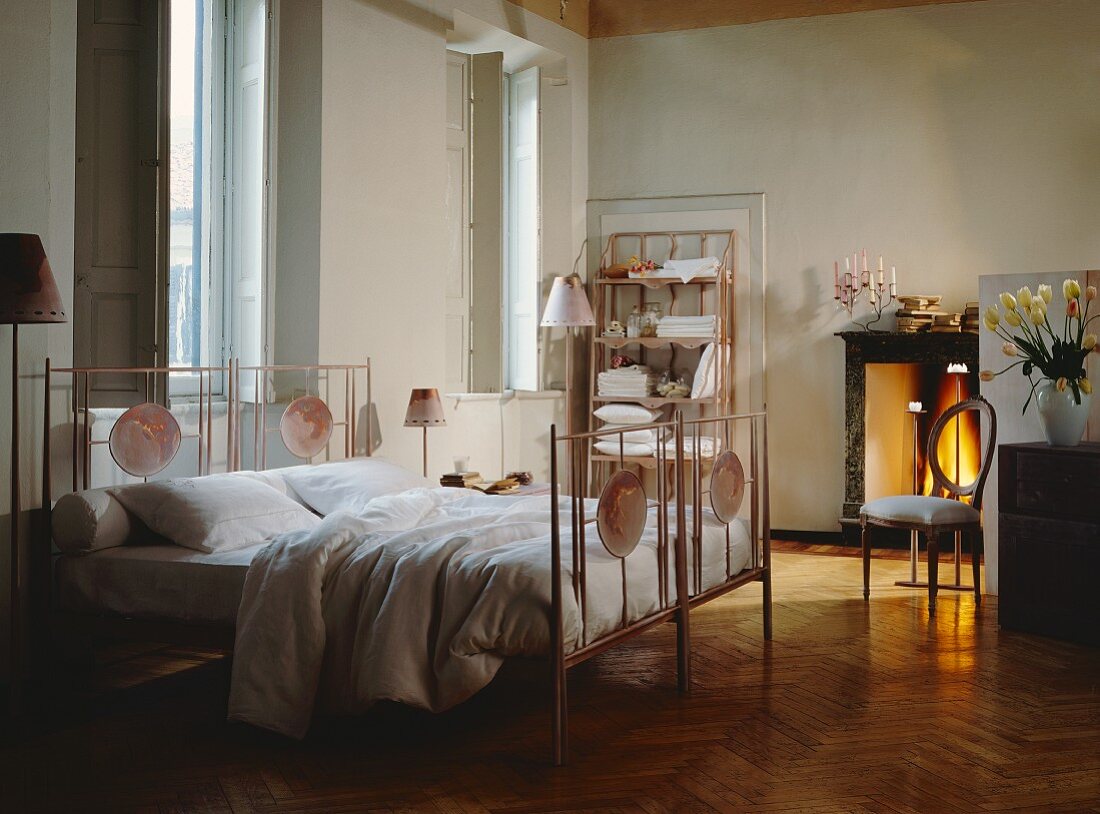 Traditional bedroom with tall windows and large metal bed with painted oval panels; firelight from open fireplace in background