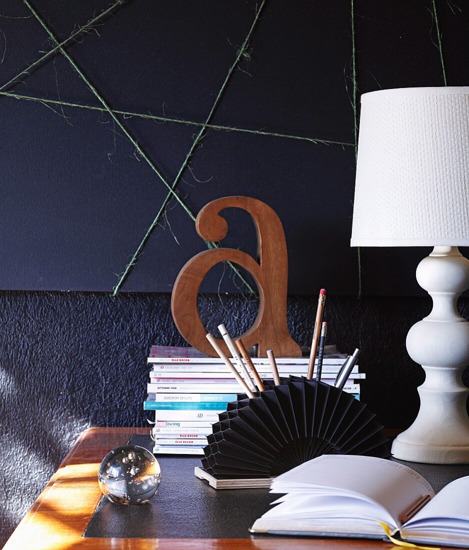 Wooden letter on stack of magazines, fan-shaped pen holder and country-house-style lamp on desk against wall decorated in black