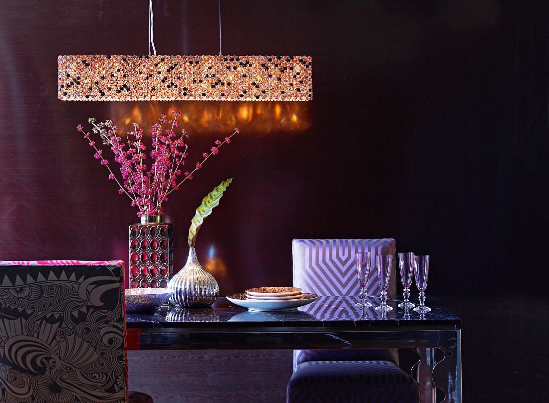 Beleuchtete Kristallpendelleuchte und eleganter Esstisch mit Champangergläsern, silberglänzender Vase und rosafarbenen Blütenzweigen