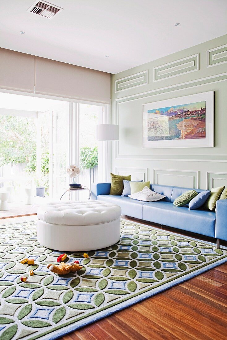 weiße Ottomane auf grafisch gemustertem Teppich und blaue Oversize-Couch vor klassischer, pastellgrüner Holzvertäfelung