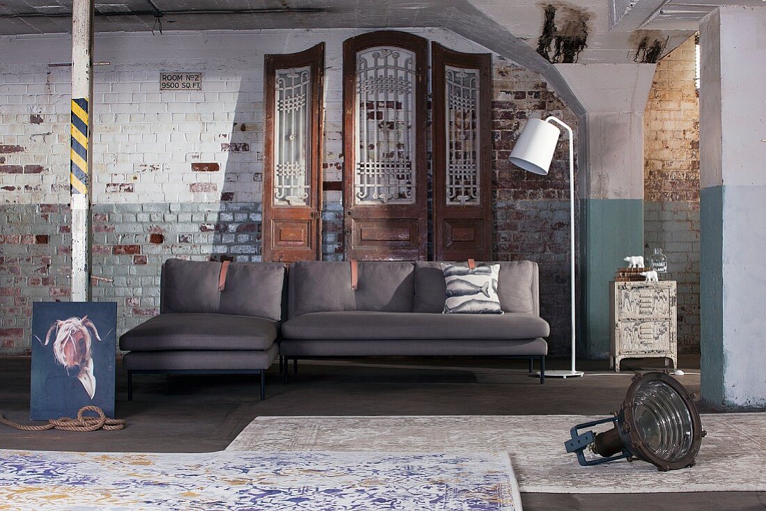 Absurdist arrangement of chaise sofa, standard lamp and rugs against old brick wall
