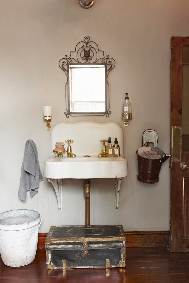 Wrought iron mirror above old stone sink with perfume bottles: enamel bucket of fresh towels hung on wall and antique trunk