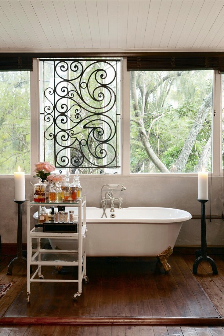 Badewanne mit Klauenfüssen aus Messing vor Fensterfront mit Schmiedeeisengitter; Rollregal mit Badeutensilien