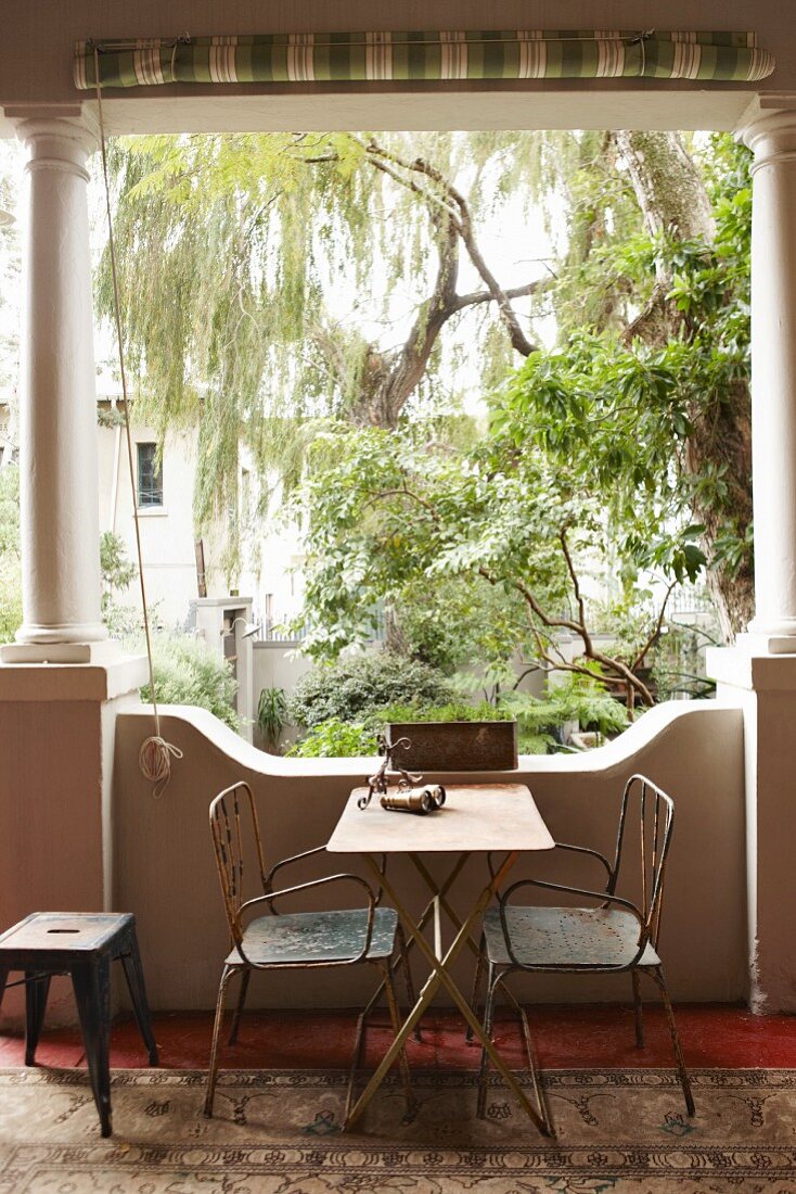 Old metal chairs and metal table in loggia with antique stone columns; view into lush courtyard