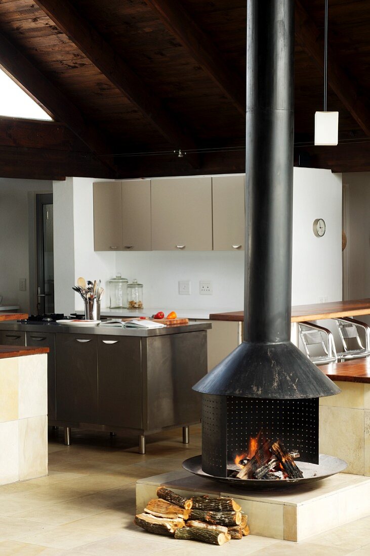 Wood-burning stove in open-plan interior in front of kitchen area with island counter and grey fitted units
