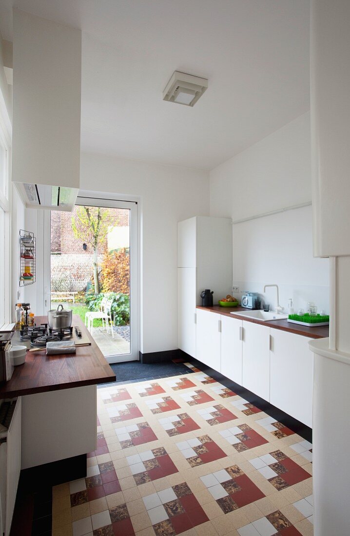 Modern, white fitted kitchen with unusual pattern of floor tiles and wide glass door leading to garden