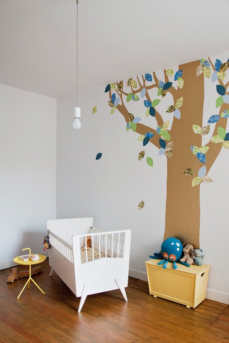 Kinderzimmer mit Babybett vor einem auf die Wand aufgemalten Baumstamm mit stilisierten Blättern