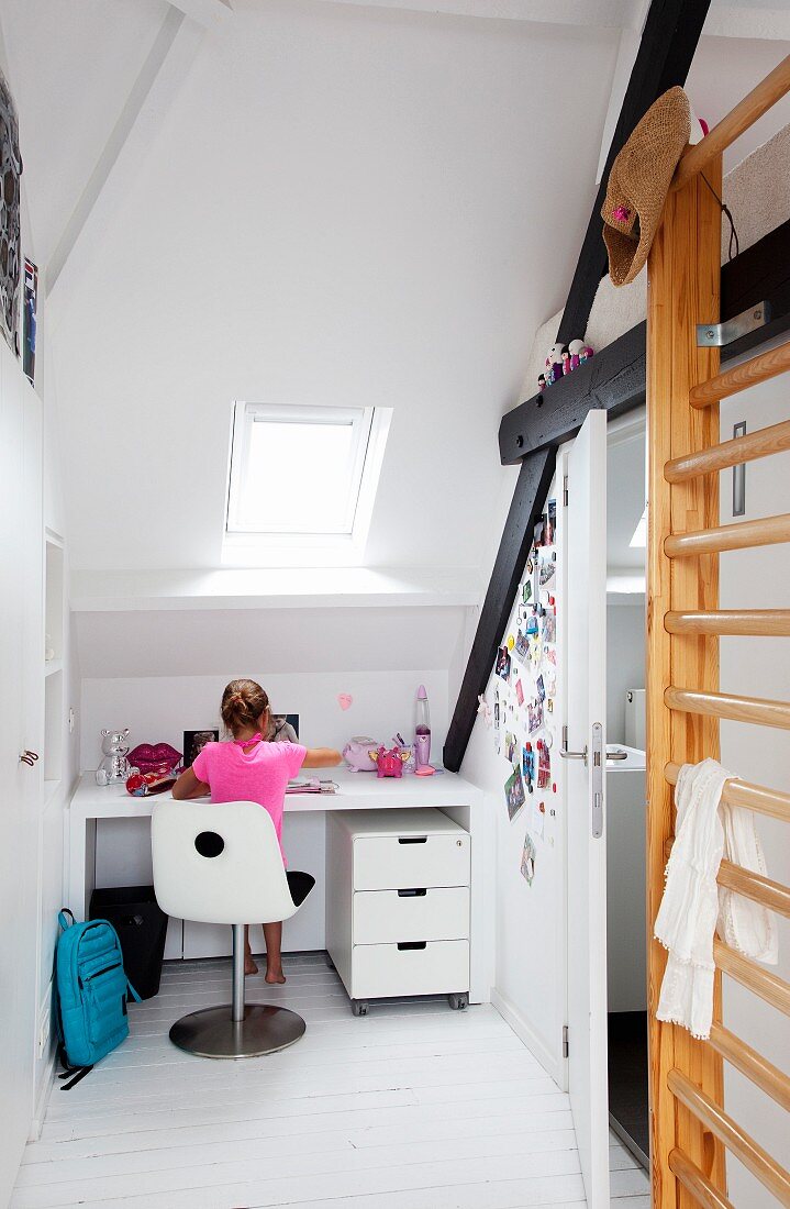 White, child's bedroom in attic - child sitting on swivel chair at fitted desk below sloping ceiling