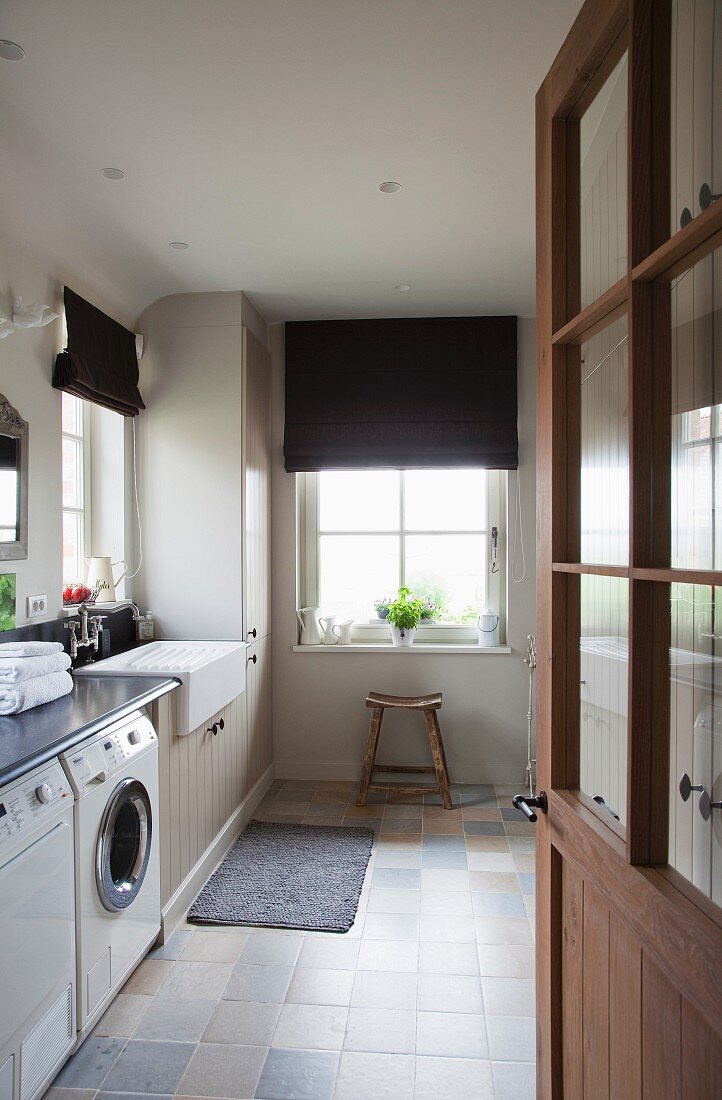 Blick in Hausarbeitsraum mit Waschmaschine, dunklen Faltrollos und Fliesen in hellen Naturtönen
