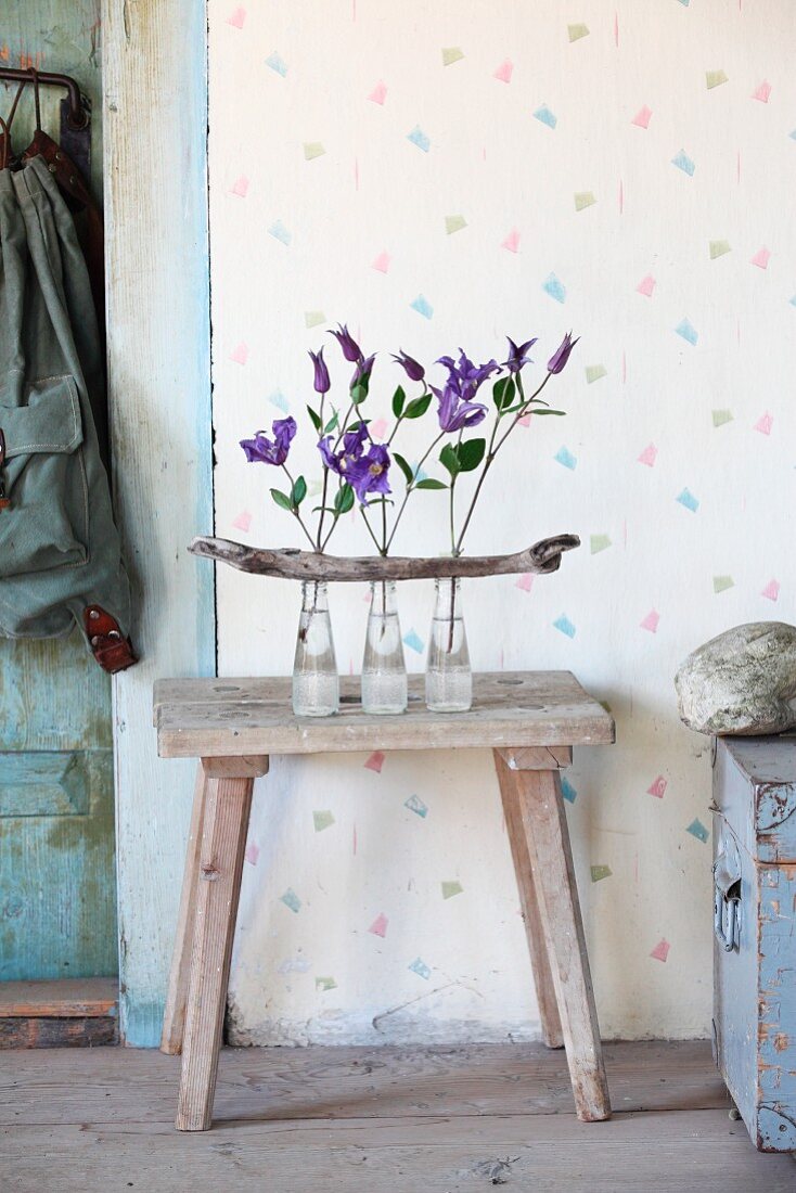 Clematisblüten in drei Glasfläschen mit Treibholzast arrangiert auf Holzhocker in Vintageambiente
