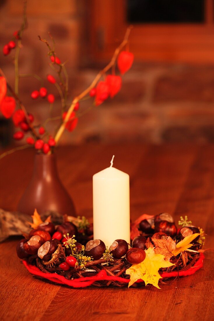 weiße Kerze in Kranzmitte aus Kastanien und Herbstblättern vor Vase mit Hagebuttenzweigen auf Tisch