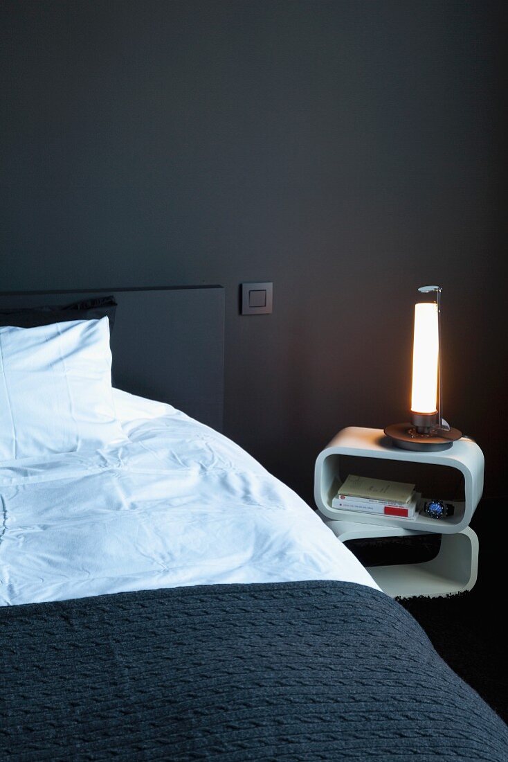 Bett mit schwarzer Tagesdecke neben Retro Nachttisch und leuchtender Tischlampe in schwarz getöntem Schlafzimmer