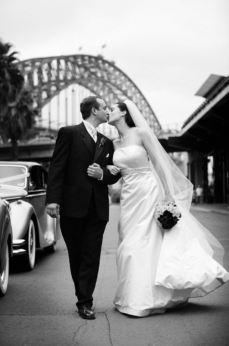 Braut in langem weißem Kleid und Bräutigam in schwarzem Anzug küssen sich auf der Straße (s-w-Aufnahme)