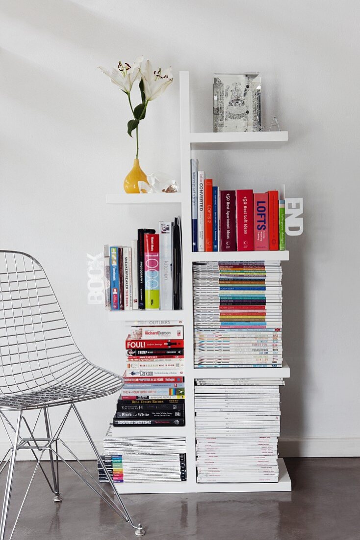 Bücher und Zeitschriften auf Designerregal mit auskragenden Fächern, seitlich ein Drahtgitterstuhl von Eames