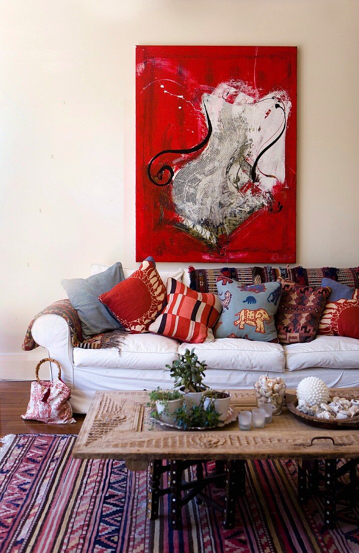 Moderne Malerei in kräftigem Rot über Landhaus Sofa mit fröhlicher Kissensammlung; Couchtisch aus geschnitzter, umgenutzter Schranktür
