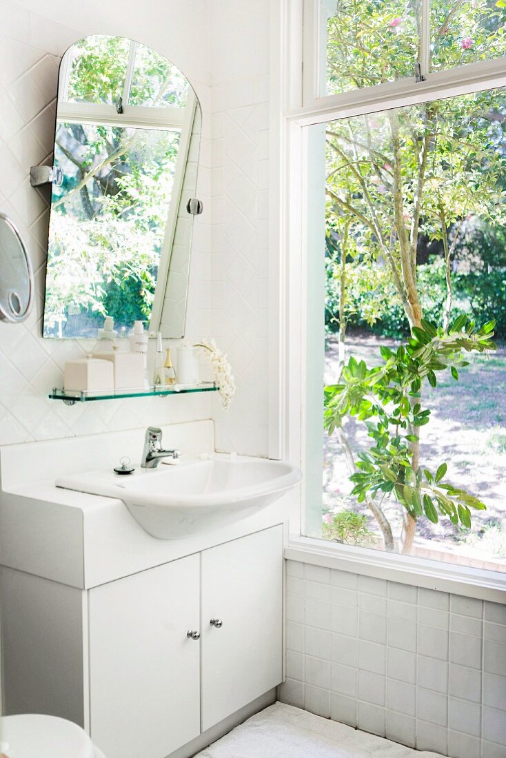 Simple, white washstand and angled mirror next to large window with view of garden