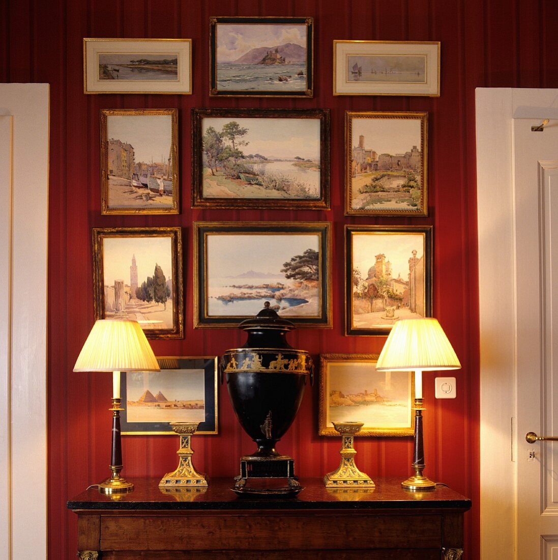 Urn-shaped vase flanked by table lamps with pleated fabric lampshades on antique cabinet below gallery of framed pictures