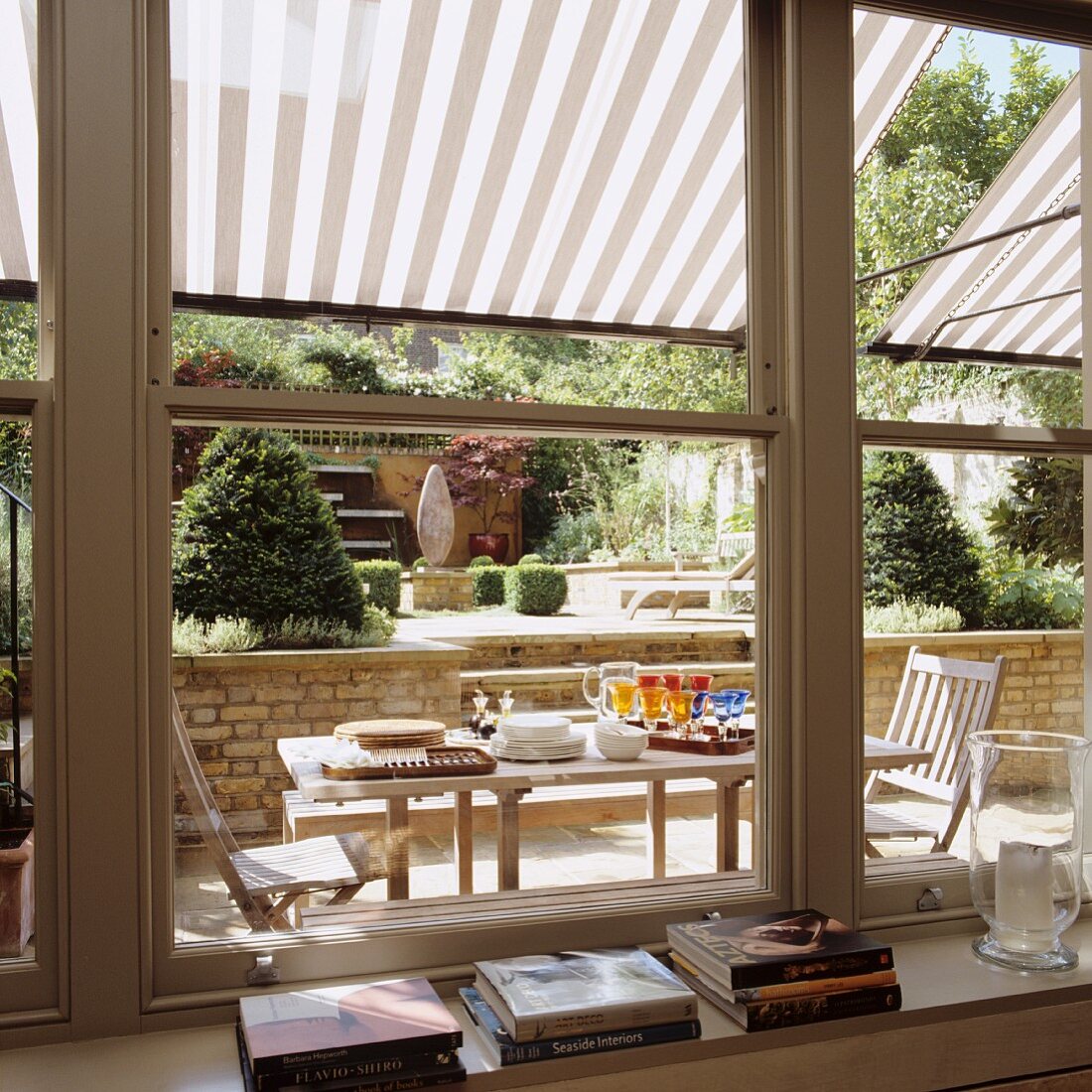 Blick durch Schiebefenster auf Terrasse mit gedecktem Tisch unter gestreifter Pergola