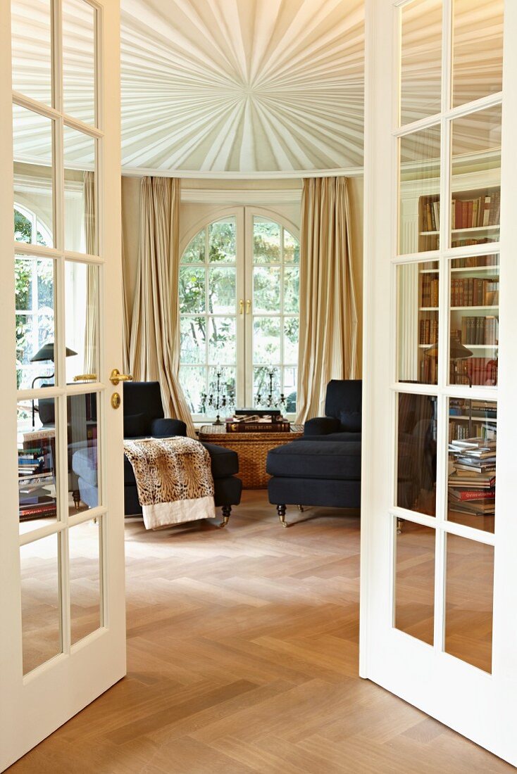 Blick durch offene Sprossentüren auf Chaiselongue am Fenster und abgehängte Decke mit strahlenförmigem Design in rundem herrschaftlichem Salon