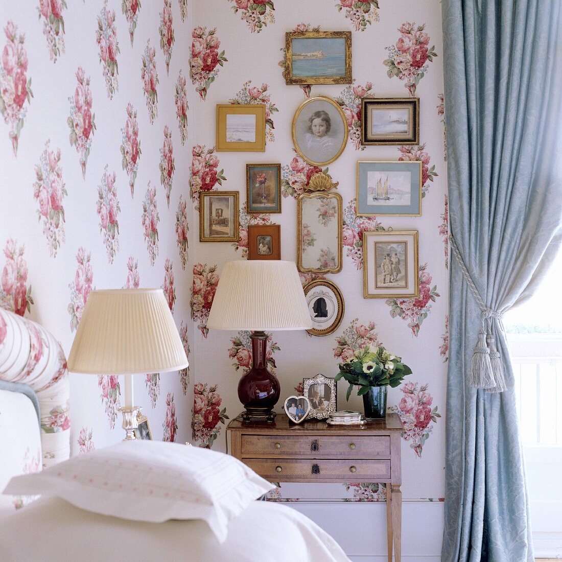 Schlafzimmerecke mit Tischleuchten und Bildersammlung vor stoffbezogener Wand mit Blumenmuster