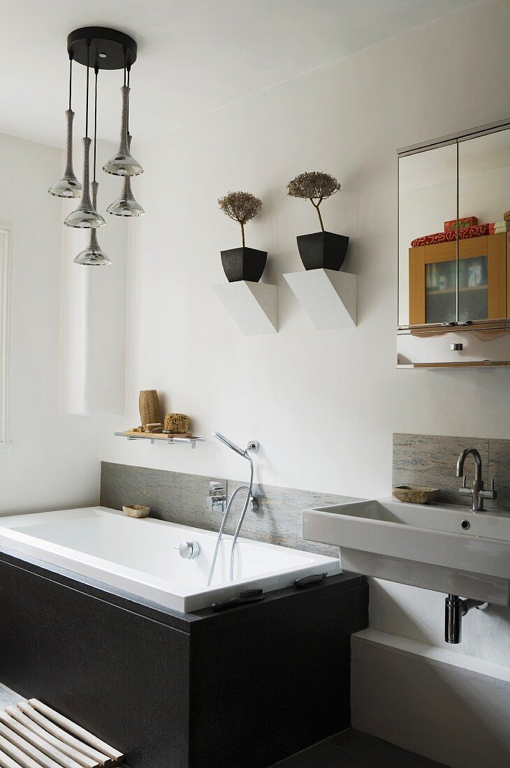 Fitted bathtub with black, stone cladding below potted plants on masonry wall brackets and next to washstand and mirrored cabinet