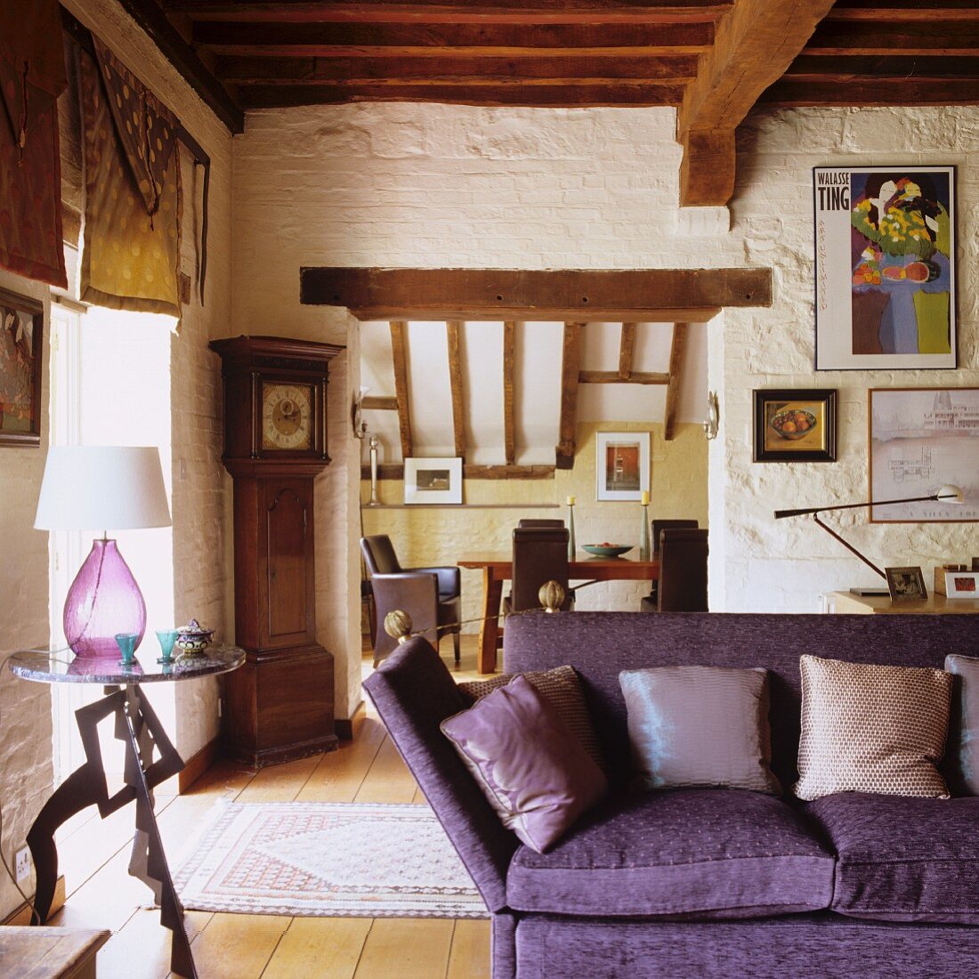 Violettes Sofa mit Kissen neben Beistelltisch in rustikalem Wohnraum mit Holzbalkendecke und Blick durch breiten Durchgang auf Essplatz