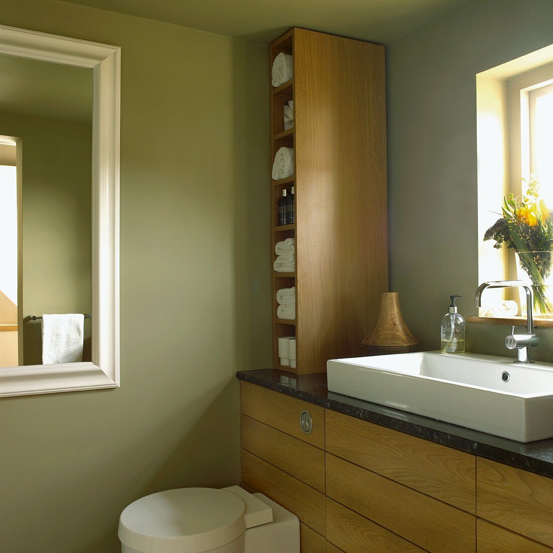 Badezimmer mit grünen Wänden, Holzmöbeln, Waschbecken vor dem Fenster, WC und Wandspiegel