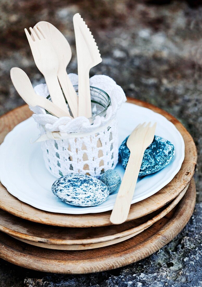 Wooden cutlery in glass jar covered in lace doily and pebbles on plate on stack of wooden boards