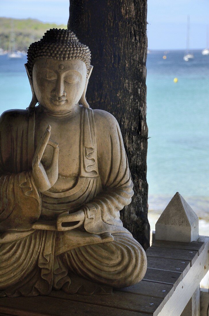 Buddhafigur aus Stein auf Holzablage am Strand mit Meerblick