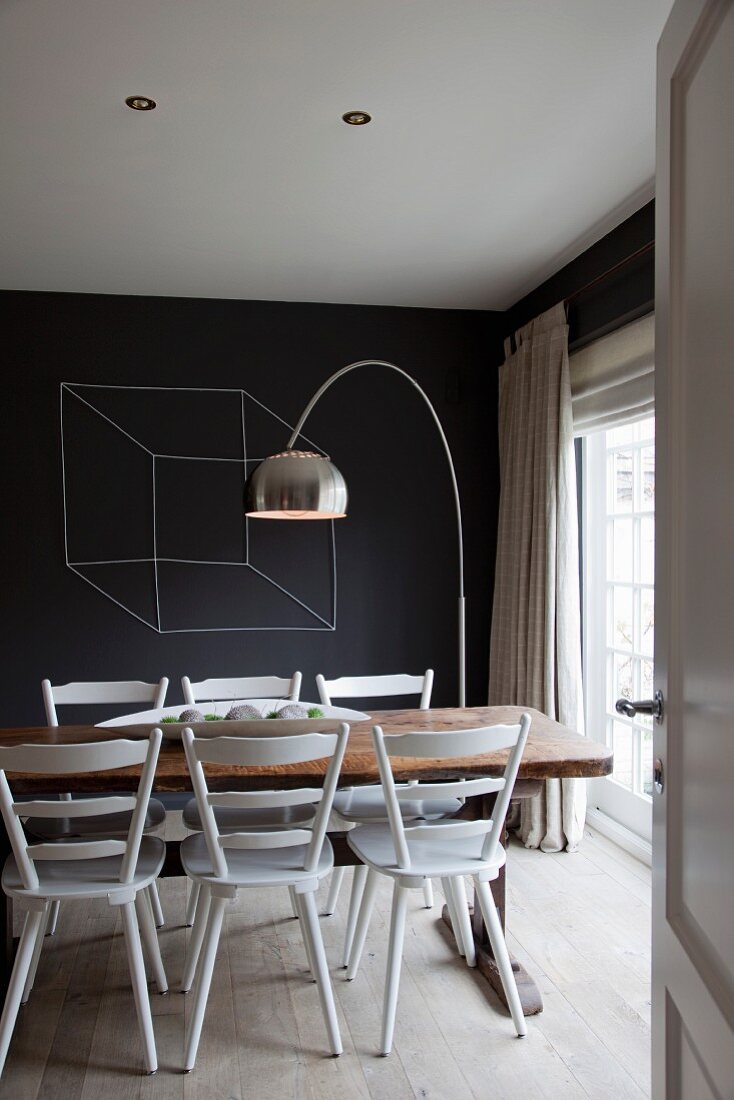 View through open door of white-painted kitchen chairs, rustic table, arc lamp and black-painted wall with drawing of cube