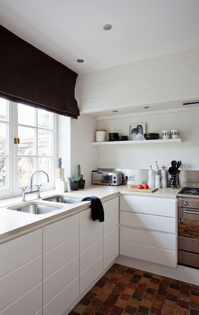 Corner of white, modern kitchen with white cupboards and integrated sinks below window with black Roman blind