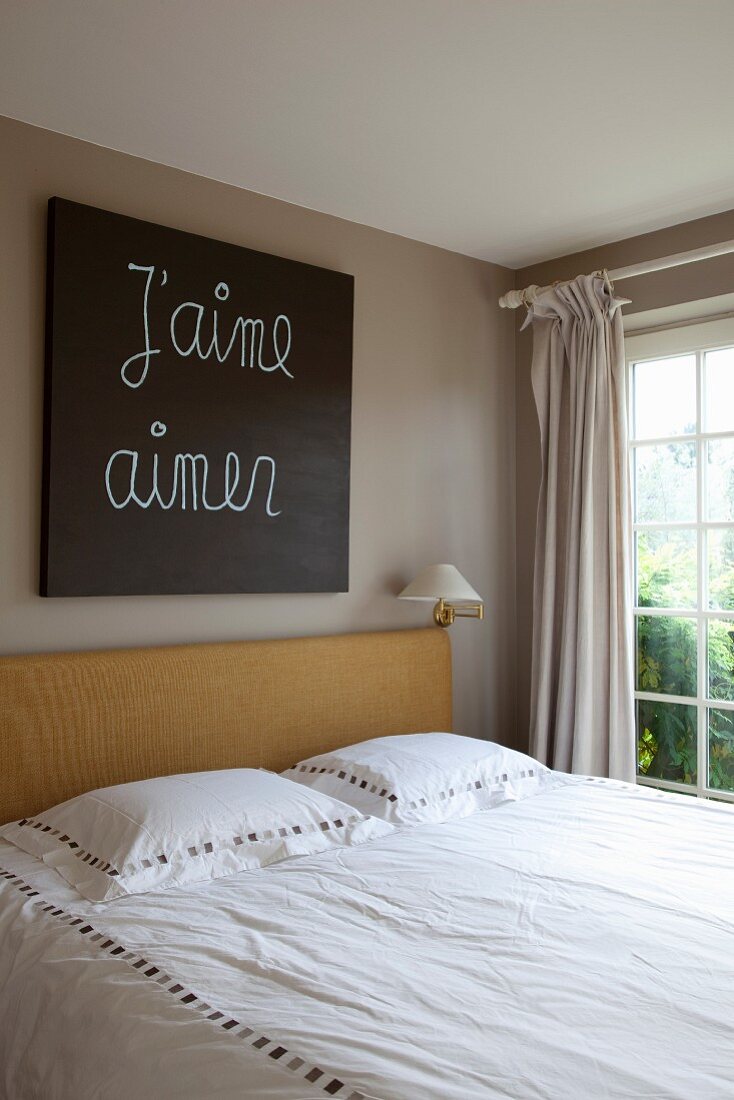 Double bed with wooden headboard below artwork with written message on wall painted pale grey