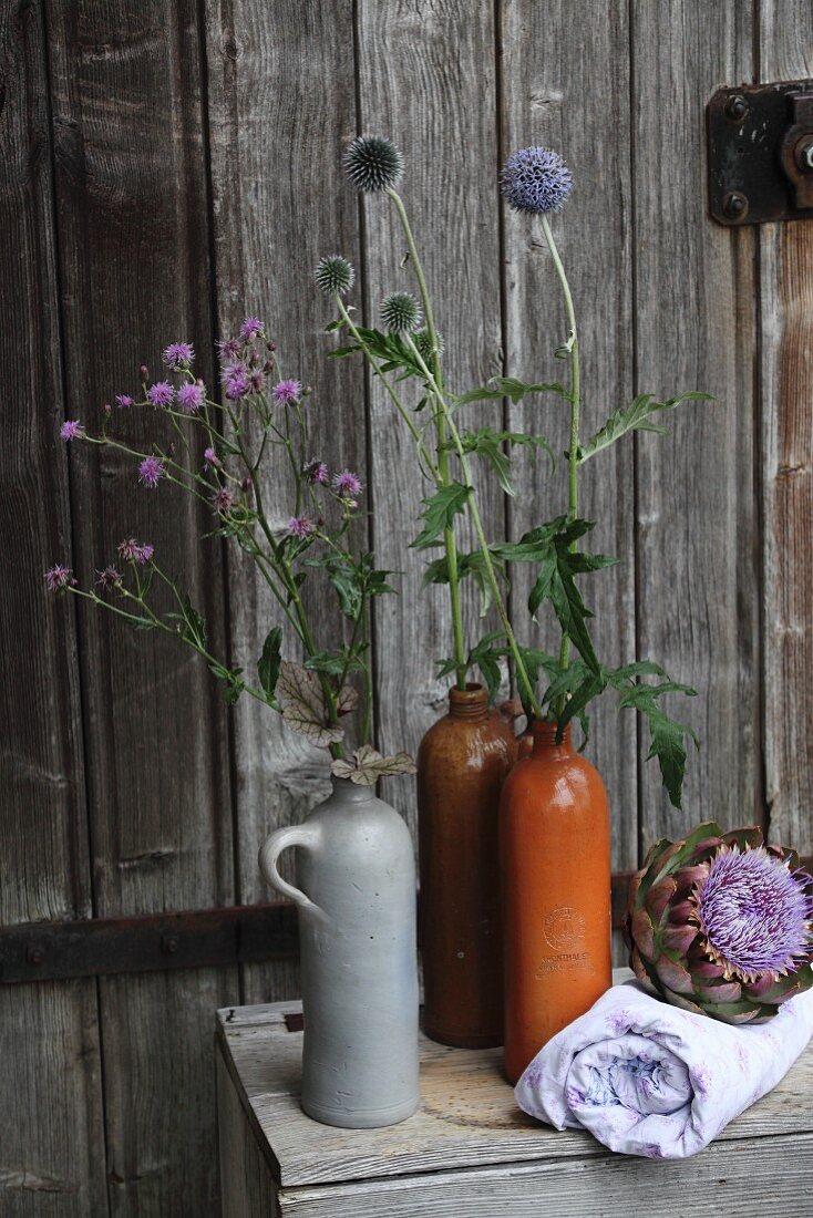 Milk thistles and globe thistles in rustic stoneware bottles and artichoke flowers arranged on pale blue cloth in front of old barn door