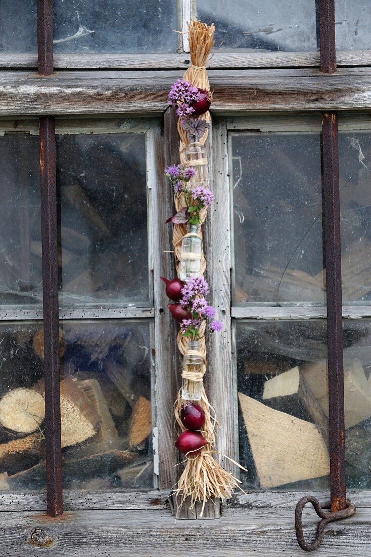 Rote Zwiebeln, Majoranblüten und mit Draht befestigte Fläschchen an Zopf aus Naturbast, aufgehängt an altem Scheunenfenster