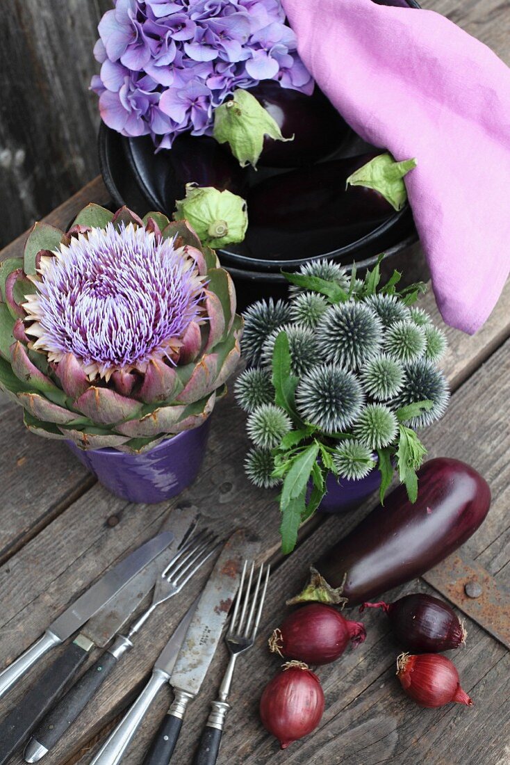 Globe thistles and artichoke flower arranged in ceramic pots next to aubergines, hydrangeas and pale violet cloth