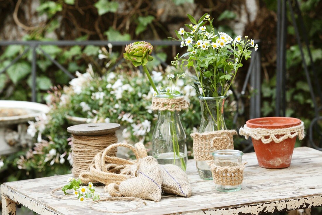Selbstgebastelte Jutesäckchen und Glasgefässe mit Strickhüllen auf rustikalem Gartentisch