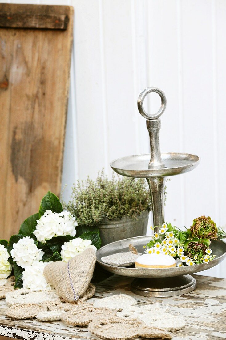 Hand-sewn hessian heart and crocheted doilies made from parcel string in front of chamomile on cake stand, white hydrangea and thyme in zinc pot