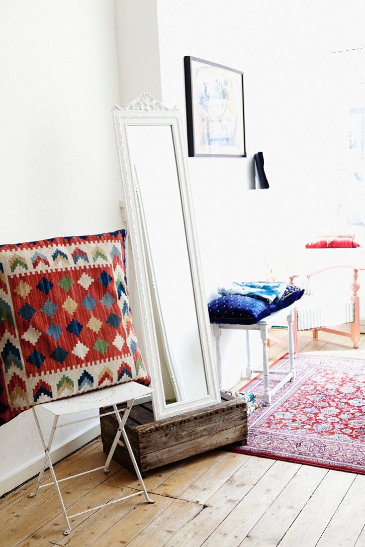 Large 'ethnic' pillow on a white metal folding chair; next to it a full length mirror leaning on a old wooden box