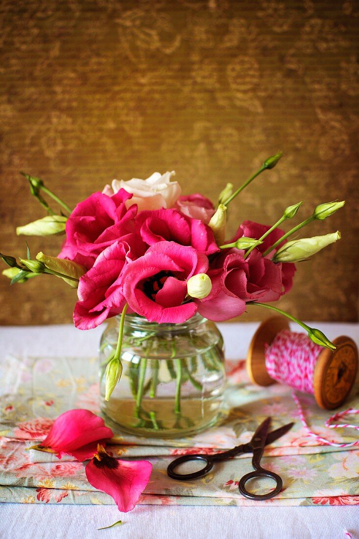 Zartes, kleines Blumensträußchen in rosa und weiß gehalten in einem Schraubverschlussglas auf blumigen gefalteten Servietten; rotweiße Kordel auf einer hölzernen Fadenrolle und einer kleinen Schere im Vintageambiente