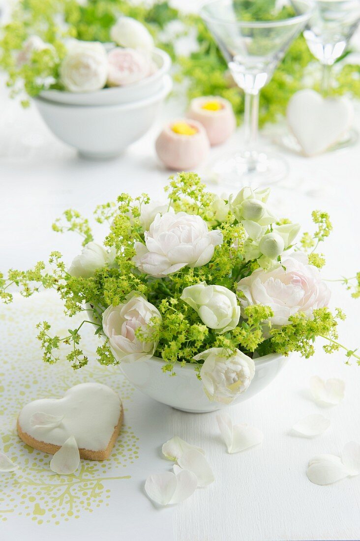 Gesteck aus weissen Rosen und Frauenmantel in Porzellanschüsseln, daneben ein Herz-Plätzchen mit Zuckerguss
