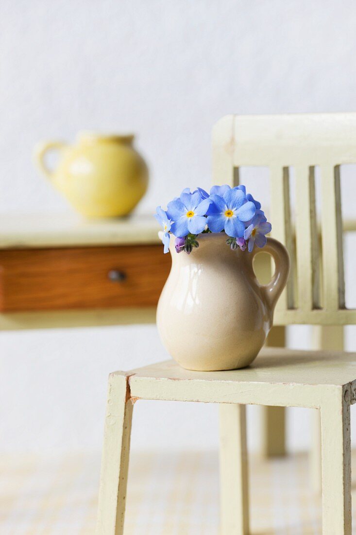 Einige Blüten Vergissmeinnicht in kleinem Krug auf Puppenstuben-Stuhl vor Holztischchen