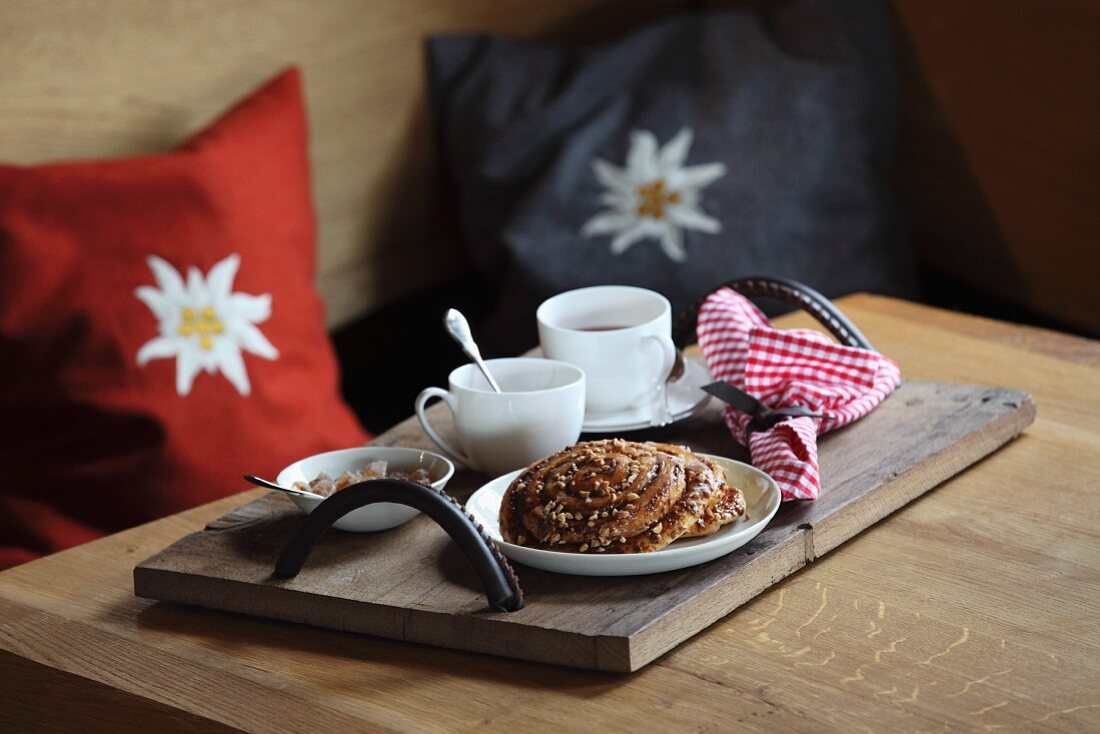 Teller mit Nussschnecken, Teetassen und Kandiszucker auf Holztablett mit selbstgemachten Ledergriffen; im Hintergrund Edelweisskissen