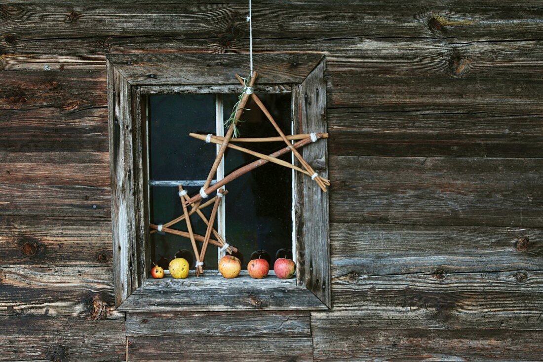 Stars hand-crafted from twigs in front of Advent arrangement of apples on window sill of wooden cabin